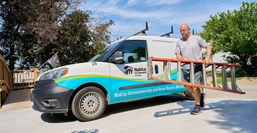 A Habitat of Omaha branded work van is parked outside a home, with a worker on a ladder repairing part of the house.