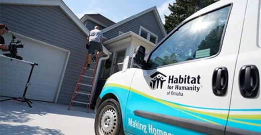 A Habitat of Omaha branded work van is parked outside a home, with a worker on a ladder repairing part of the house.