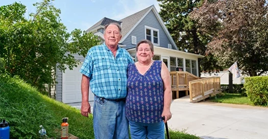 A senior couple poses in front of their house, smiling after a Habitat home repair of their front porch and driveway to add an accessible entrance.