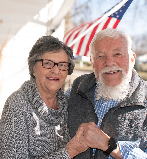 A senior couple smiles broadly and holds hands while posing in front of their home, which was repaired through the Habitat home repair program for veterans.