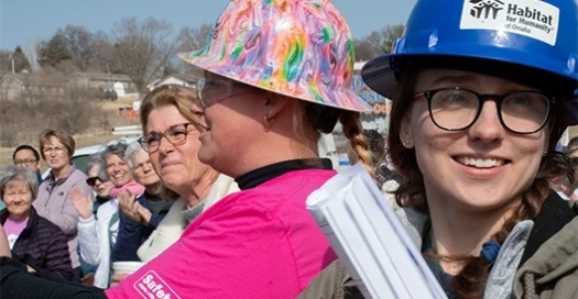 Three women in hard hats are in the foreground among a larger group of Habitat volunteers receiving on site training.