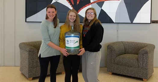 three young women pose with a water cooler jug full of spare change to donate to Habitat Omaha