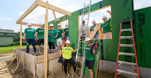 a small group of volunteers in hard hats pose near a ladder and frame of a future Habitat house.