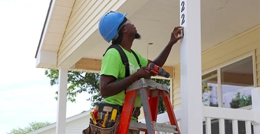 male volunteer stands on a ladder with a tool belt, add house numbers to a new Habitat home in Omaha