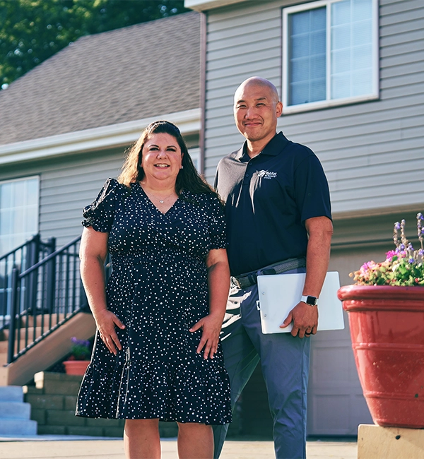 A Habitat home repair employee and a homeowner stand outside the home, smiling and posing together.