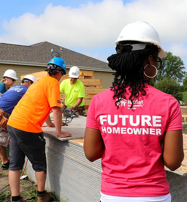 A volunteer with her back facing away from us wears a t-shirt that says “future homeowner.” She and four other volunteers wear hard hats while working on building a home with Habitat Omaha.