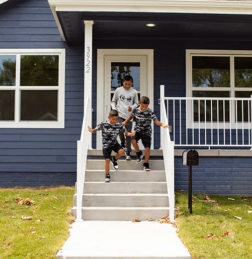 Three joyful kids run down the front steps of their newly built blue Habitat for Humanity house. The sun casts a warm glow on the scene, symbolizing hope and a brighter future.